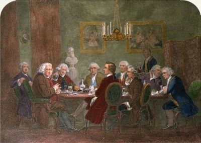 Dining room with nine men seated around a table. The dinner has been finished, and a large man at the head talks and gesticulates while the others eagerly listen. The men wear wigs and clothing of late 18th century Britain, and the furniture, hangings, and chandelier are of similar vintage. A liveried servant is entering with a tray bearing two high-shouldered decanters of wine.